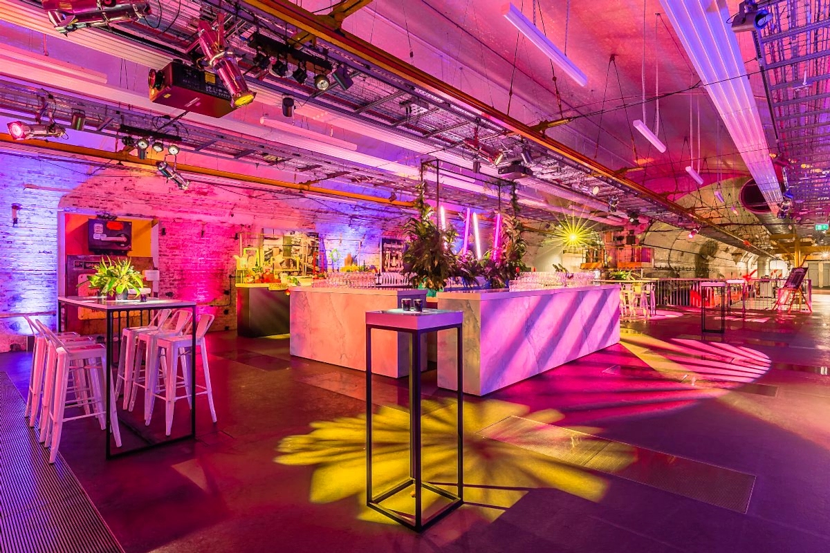 An underground venue room decorated with pink chairs, tables with flowers, and pink and yellow lighting. The Mail Rail tunnels are visible in the background.