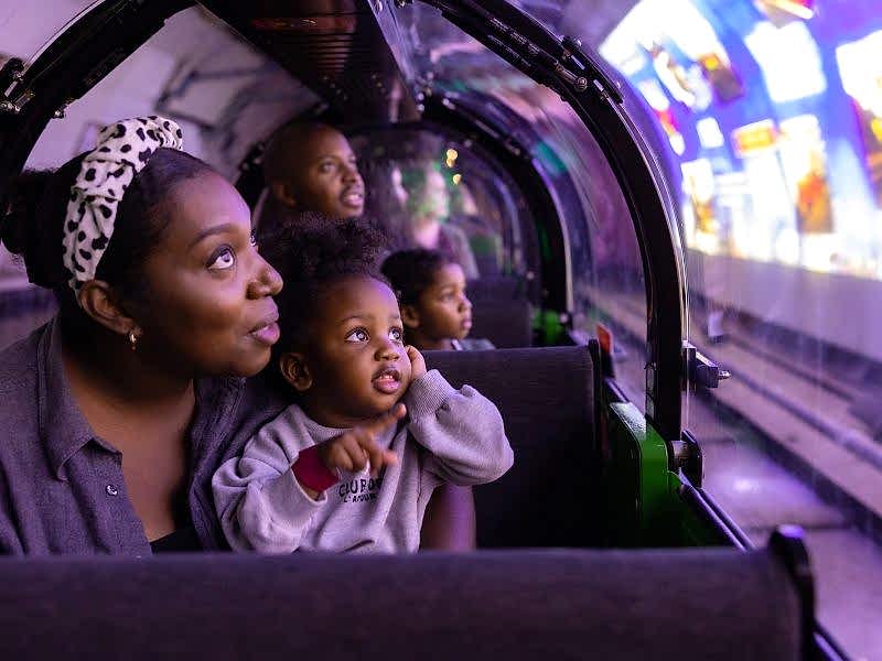 A mother and child sitting in the carriage of a miniature train. They are looking out the window at a video being projected onto the walls of the train platform.