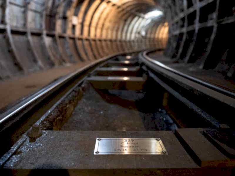 A closeup view of a silver coloured metal plaque, fixed to a wooden sleeper rail on the floor of the Mail Rail tunnel.