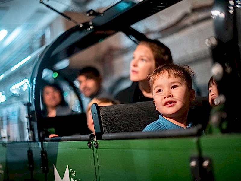A smiling young boy rides Mail Rail with his mum sat behind him. You can see the green Mail Rail carriage is full with families.