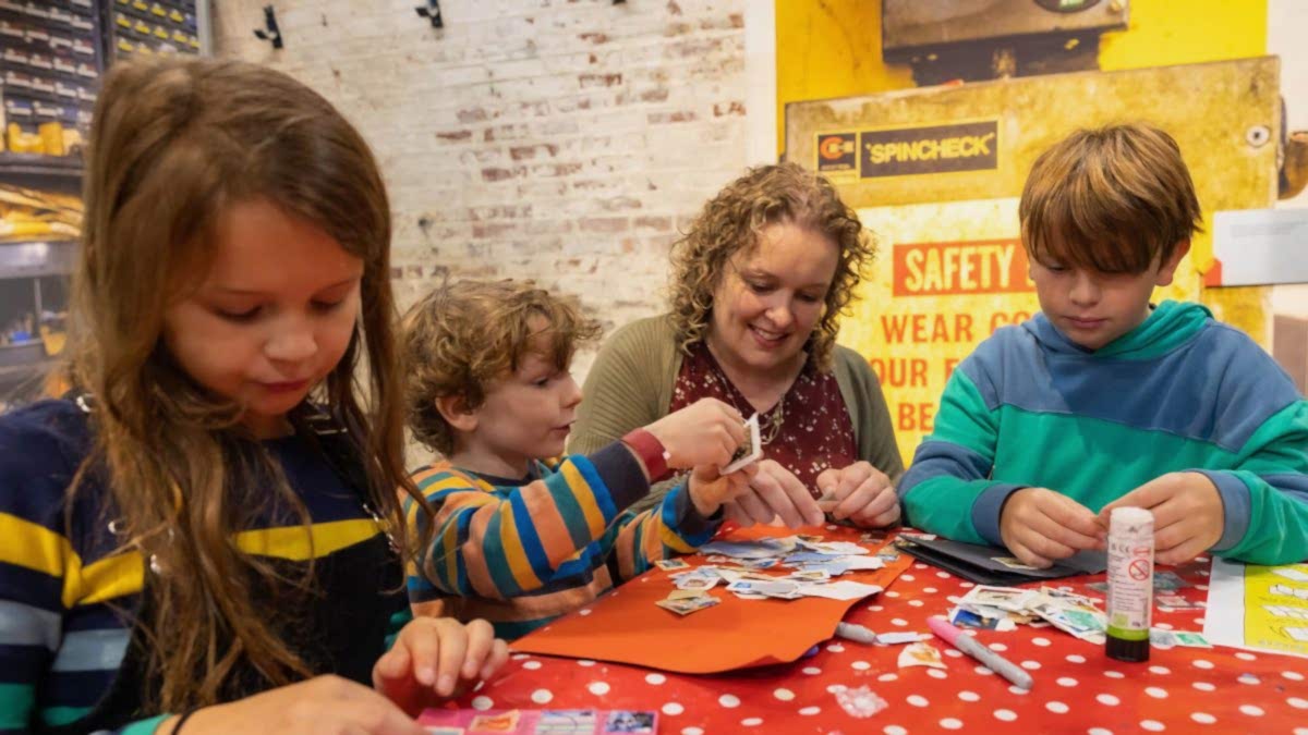A mum, with blonde curly hair, sits at her table with two fair haired boys and a browned haired girl. All of them are sticking stamps down to a piece of card.