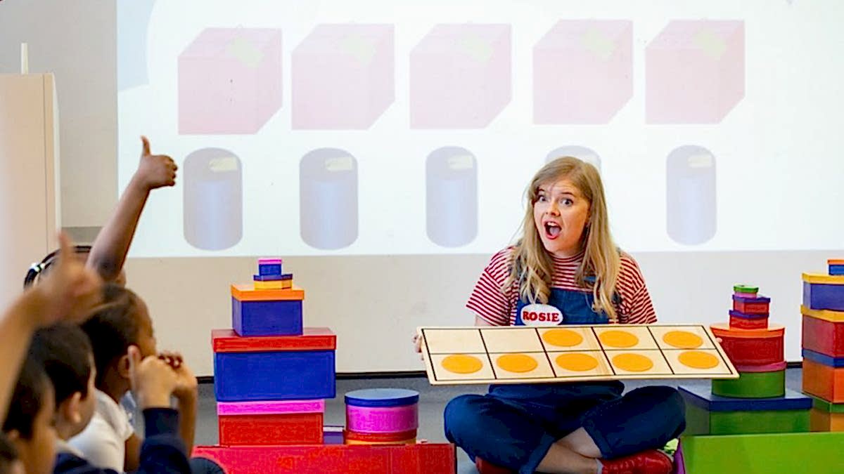 A learning facilitator is sitting on the floor surrounded by children. She holds a game and looks at them with a surprise face.