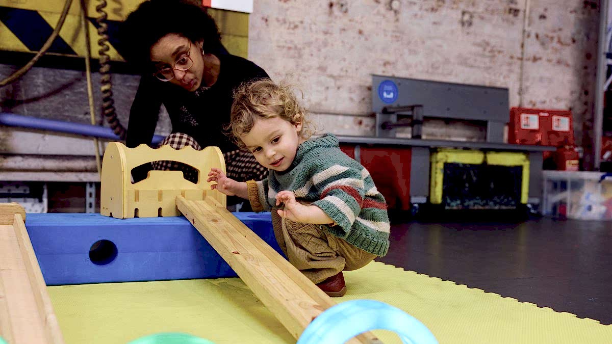 A little boy crouches by a ramp and watches his little toy car speed down it, through a colourful tunnel.
