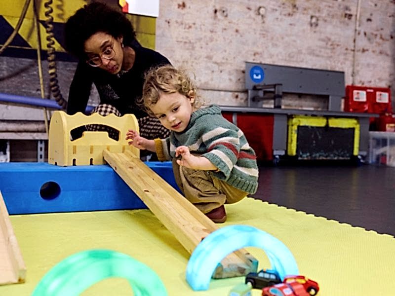 A little boy crouches by a ramp and watches his little toy car speed down it, through a colourful tunnel.
